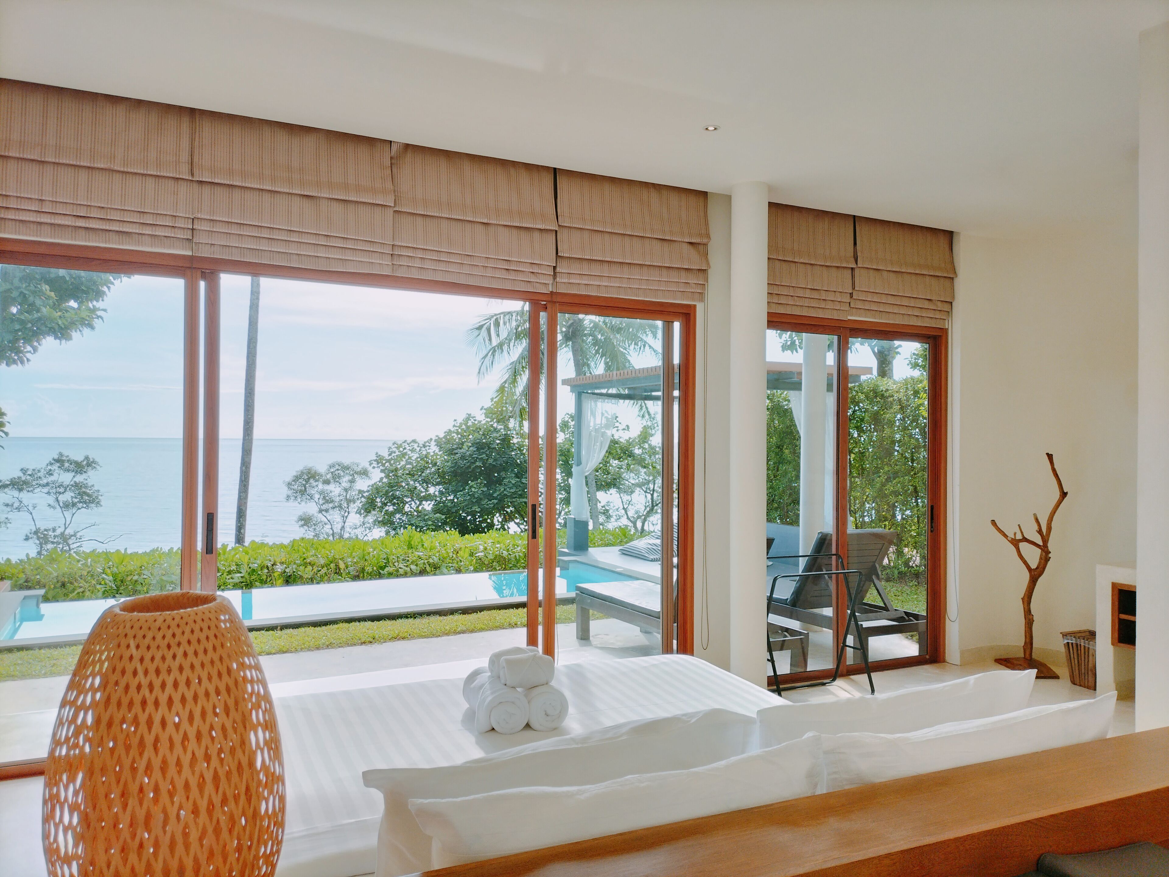 A modern hotel room with large windows, a bed, a table, and a view of the sea.