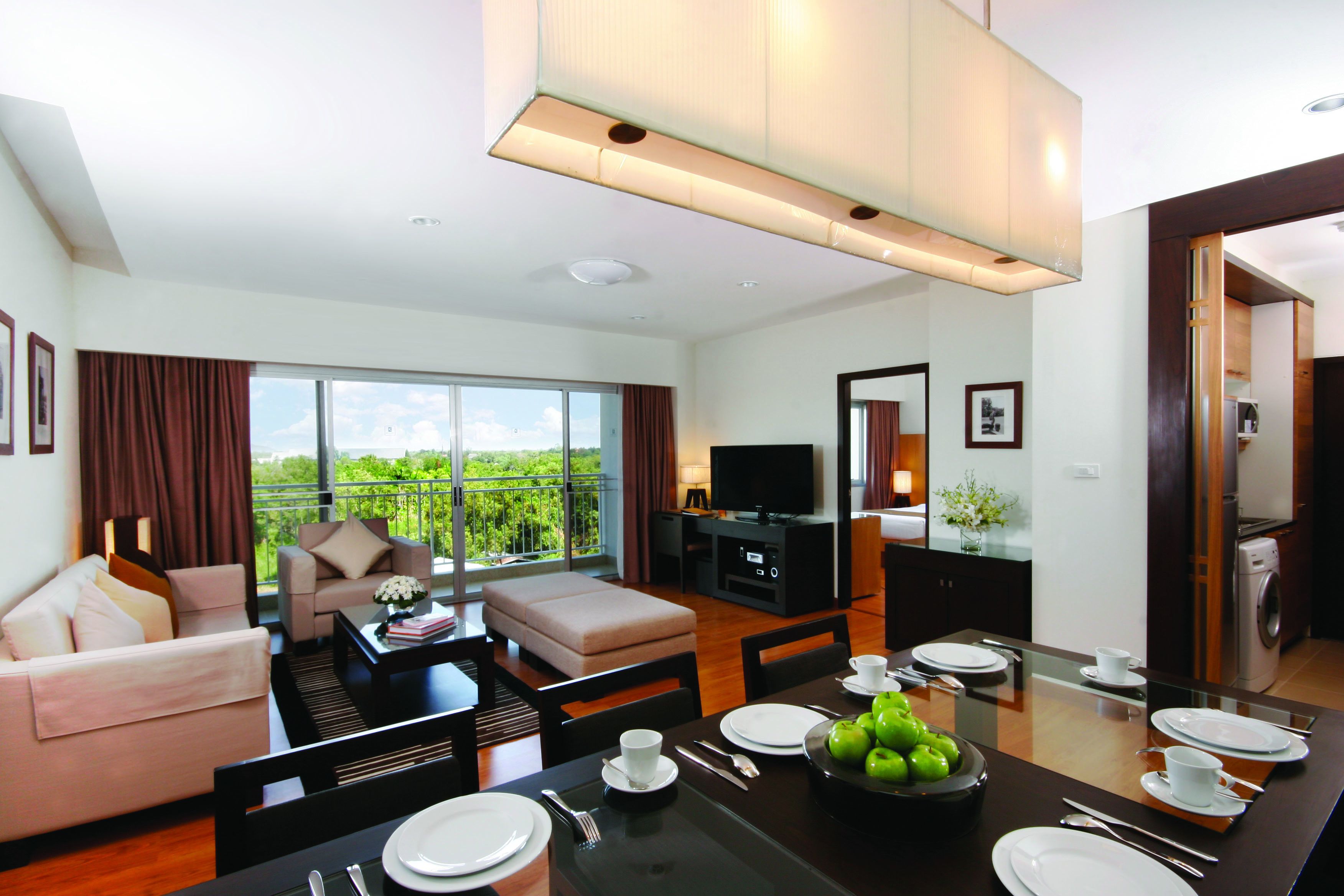 A modern hotel room with a dining area, a sofa, a TV, and a balcony with a view of the greenery.