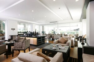 Un amplio salón con diversas opciones de asientos, una estación de café central y ventanas grandes que permiten la entrada de luz natural.