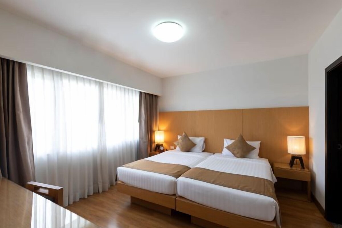 A hotel room with a double bed, two bedside tables with lamps, a wooden headboard, and a large window with curtains.