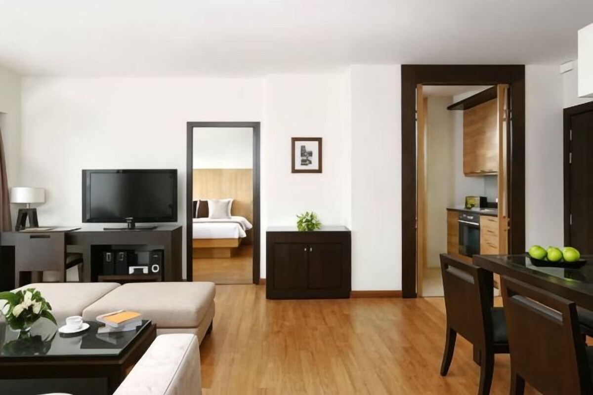 Una habitación de hotel moderna con televisión de pantalla plana, un sofá, una mesa de comedor con sillas, un mueble pequeño y una cocina visible a través de una puerta abierta.