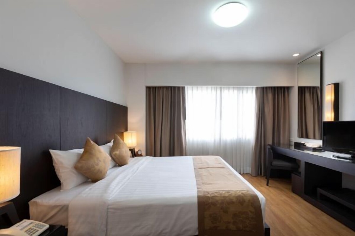 Una habitación de hotel con cama, lámparas de noche, un escritorio con televisor y un amplio ventanal con cortinas.