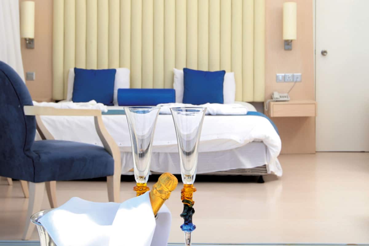 A hotel room with a bed, bedside tables, a chair, and two clear glasses on a table.