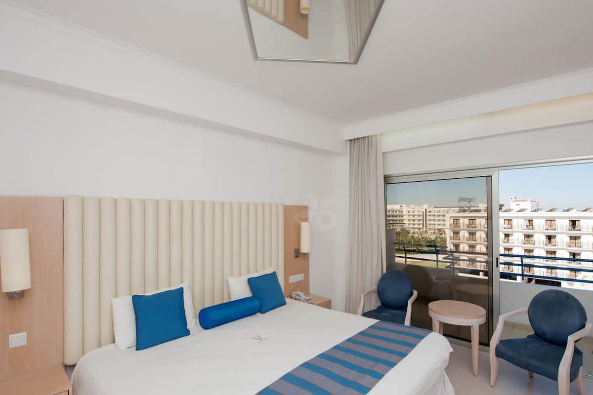 A hotel room with a large bed, a small table, two chairs, a balcony with a view of buildings, and a ceiling light fixture.