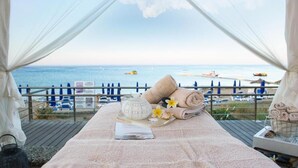 A beachside bed with towels, a tray with a glass of drink, and a view of the sea and beach umbrellas. - Vrissiana Boutique Beach Hotel (Paralimni)