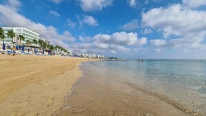 A beach with clear water, a sandy shore, and a hotel with palm trees and umbrellas. - Vrissiana Boutique Beach Hotel (Paralimni)
