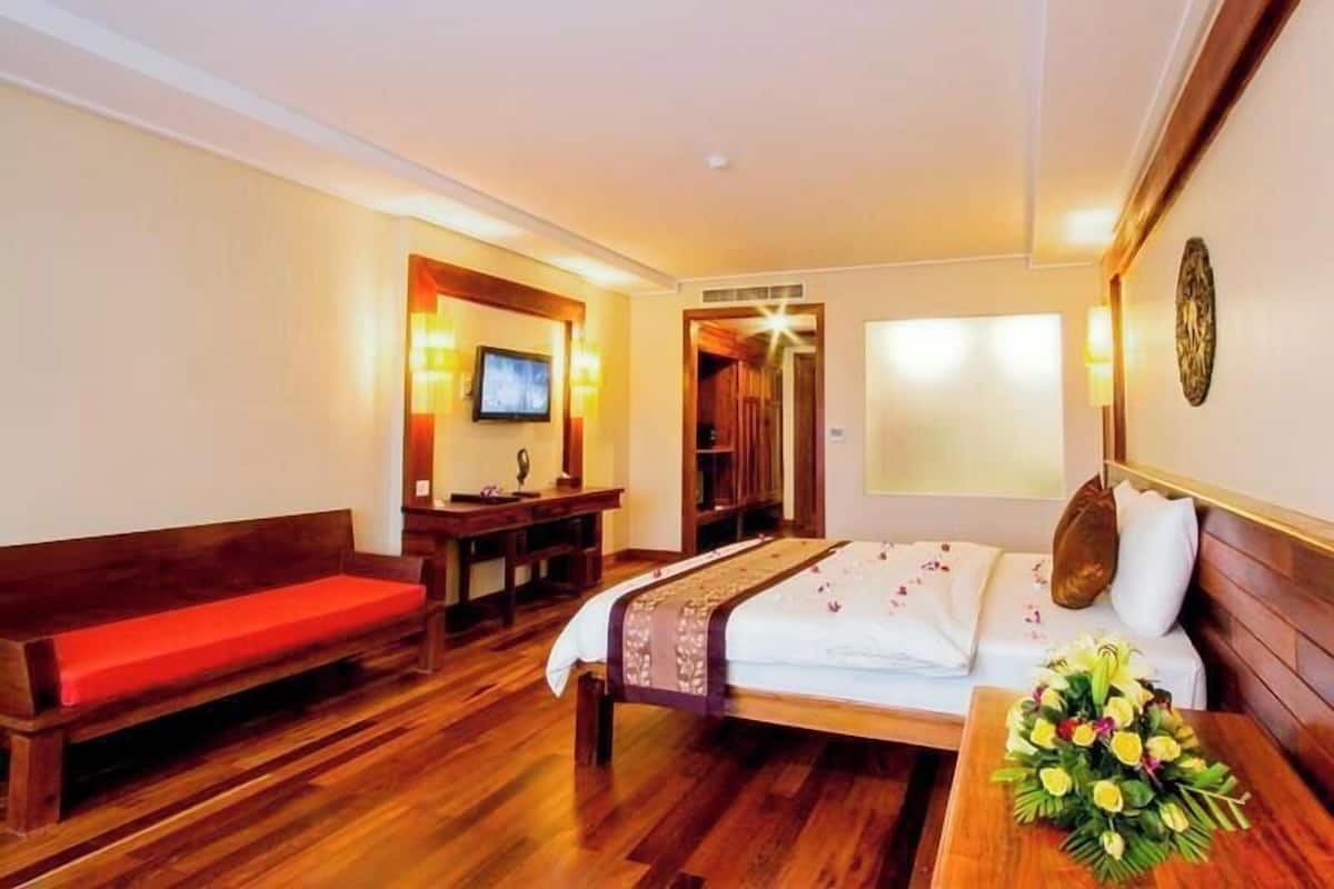 Habitación de hotel con suelo de madera, cama con cubrecama blanco, banco de madera, televisor y aire acondicionado instalado en la pared.