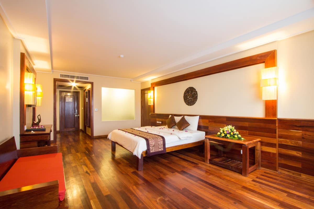 Una habitación de hotel con suelo de madera, una cama con cubrecama blanco, una mesita de noche con lámpara y un banco de madera con flores.