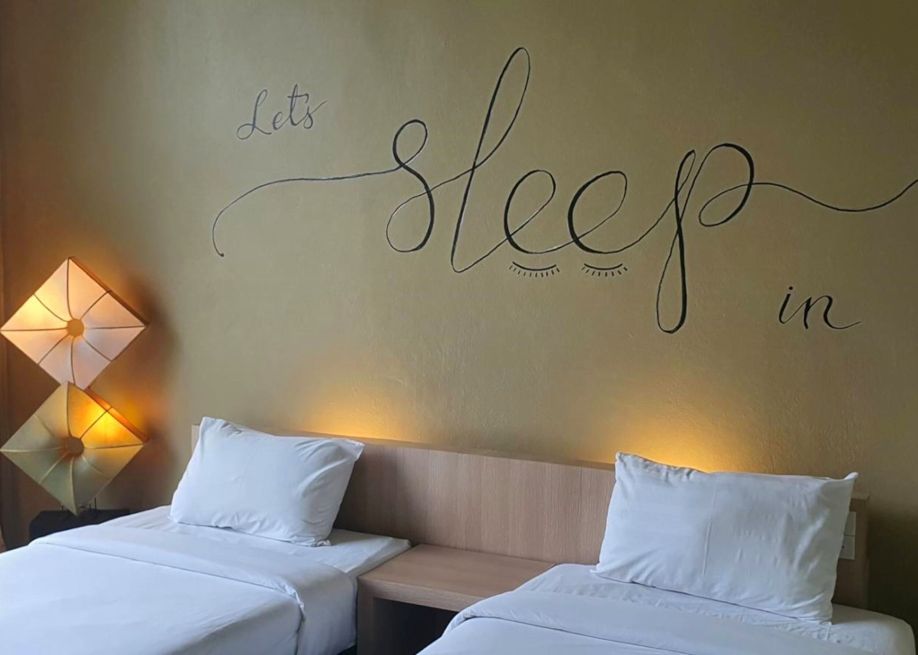 A hotel room with two beds, a wall with the phrase "Let's sleep in" and a decorative lamp.