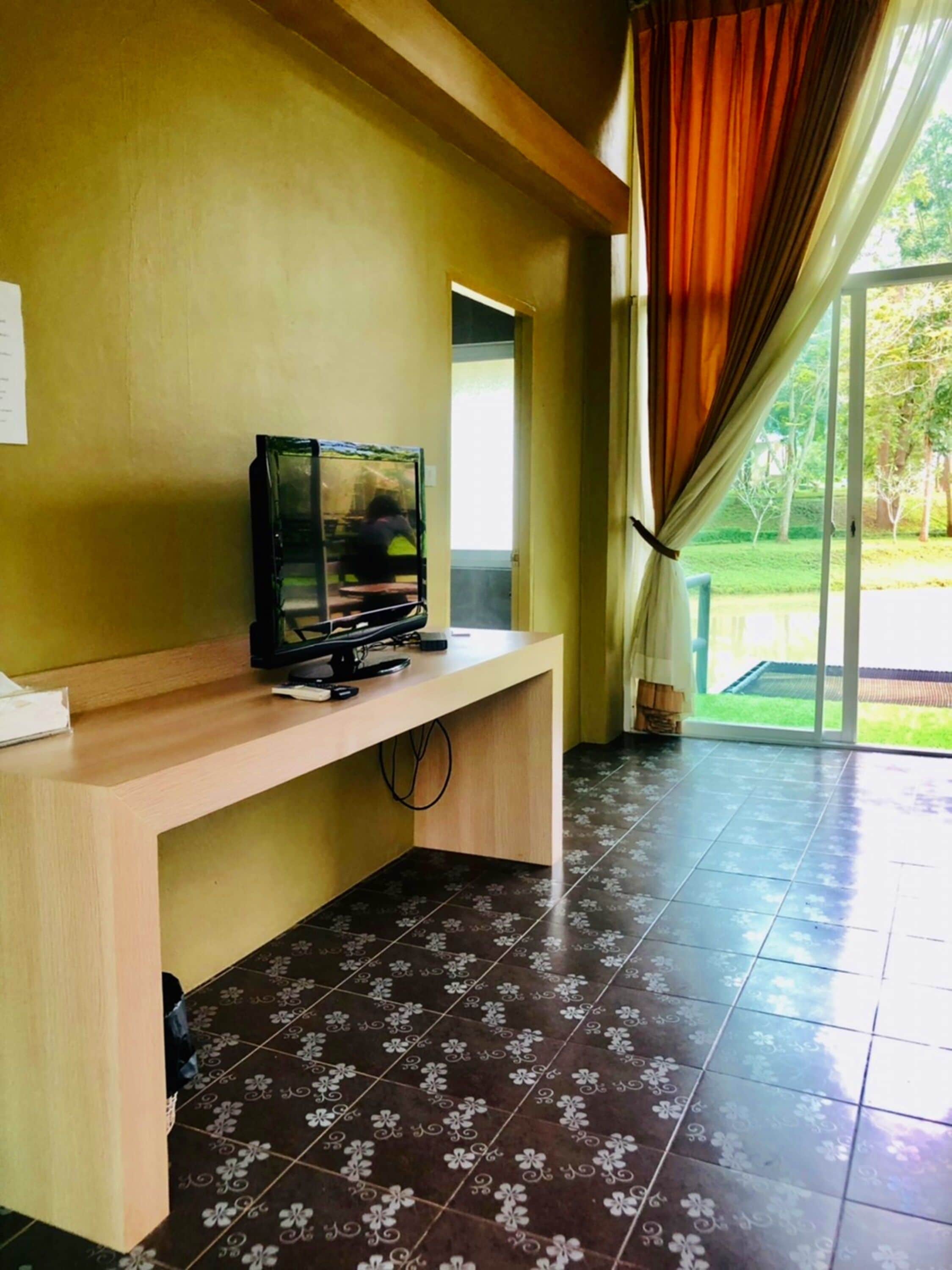 A room with a wooden desk, a television on it, and a view of a green outdoor area through a glass door.