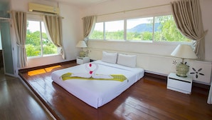 A hotel room with a large bed, two bedside tables, a window with curtains, an air conditioning unit, and a view of greenery outside.