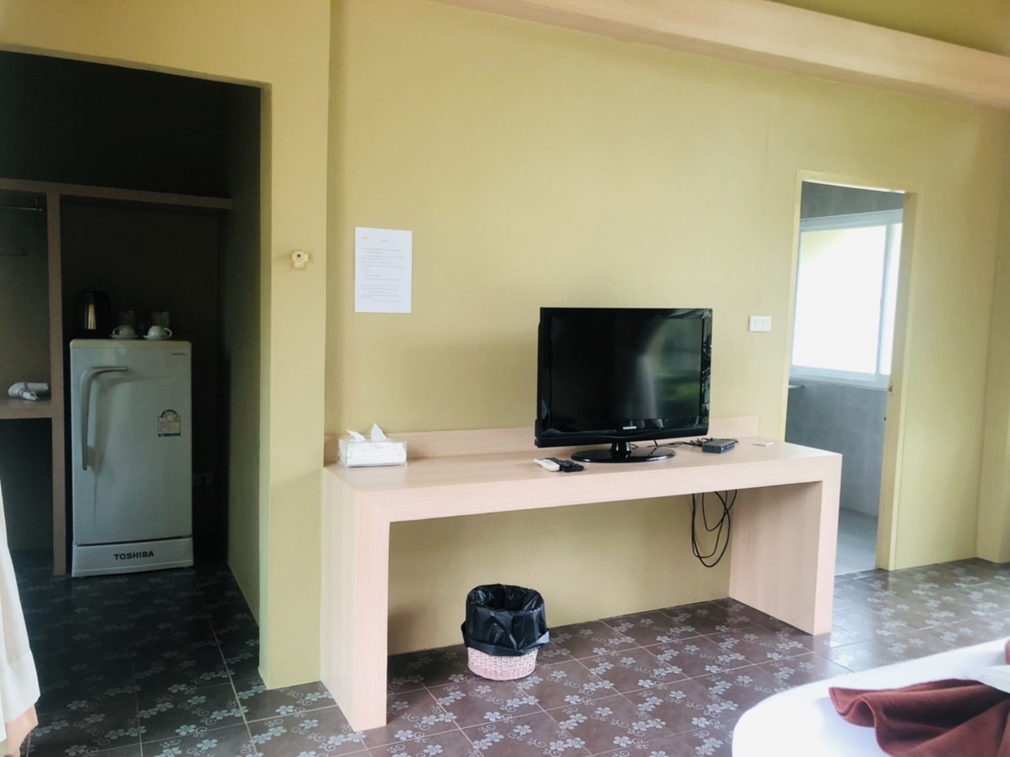 A room with a desk, a television, a refrigerator, and a trash bin.