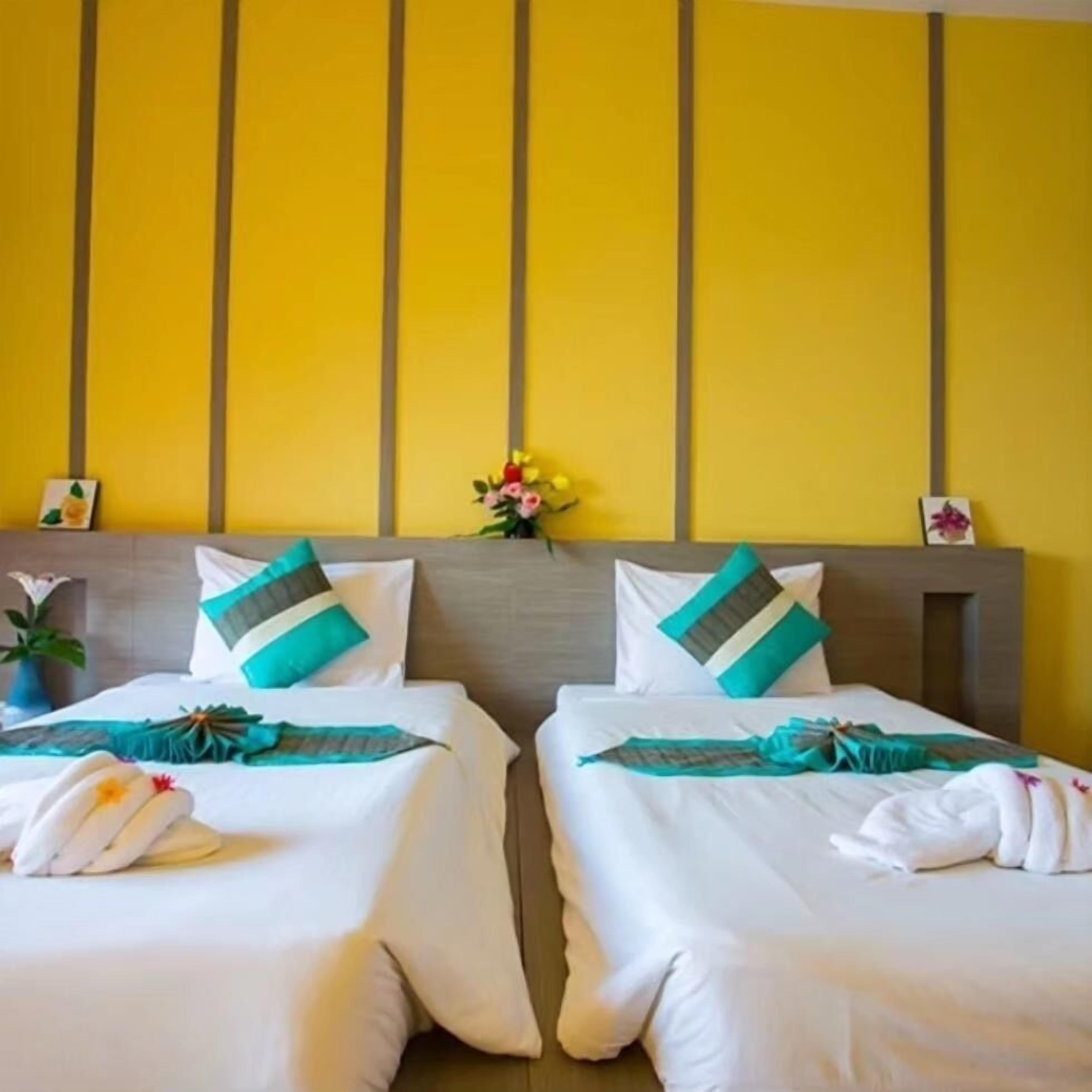 A hotel room with two beds, each with white bedding and teal pillows, against a yellow wall with vertical paneling.