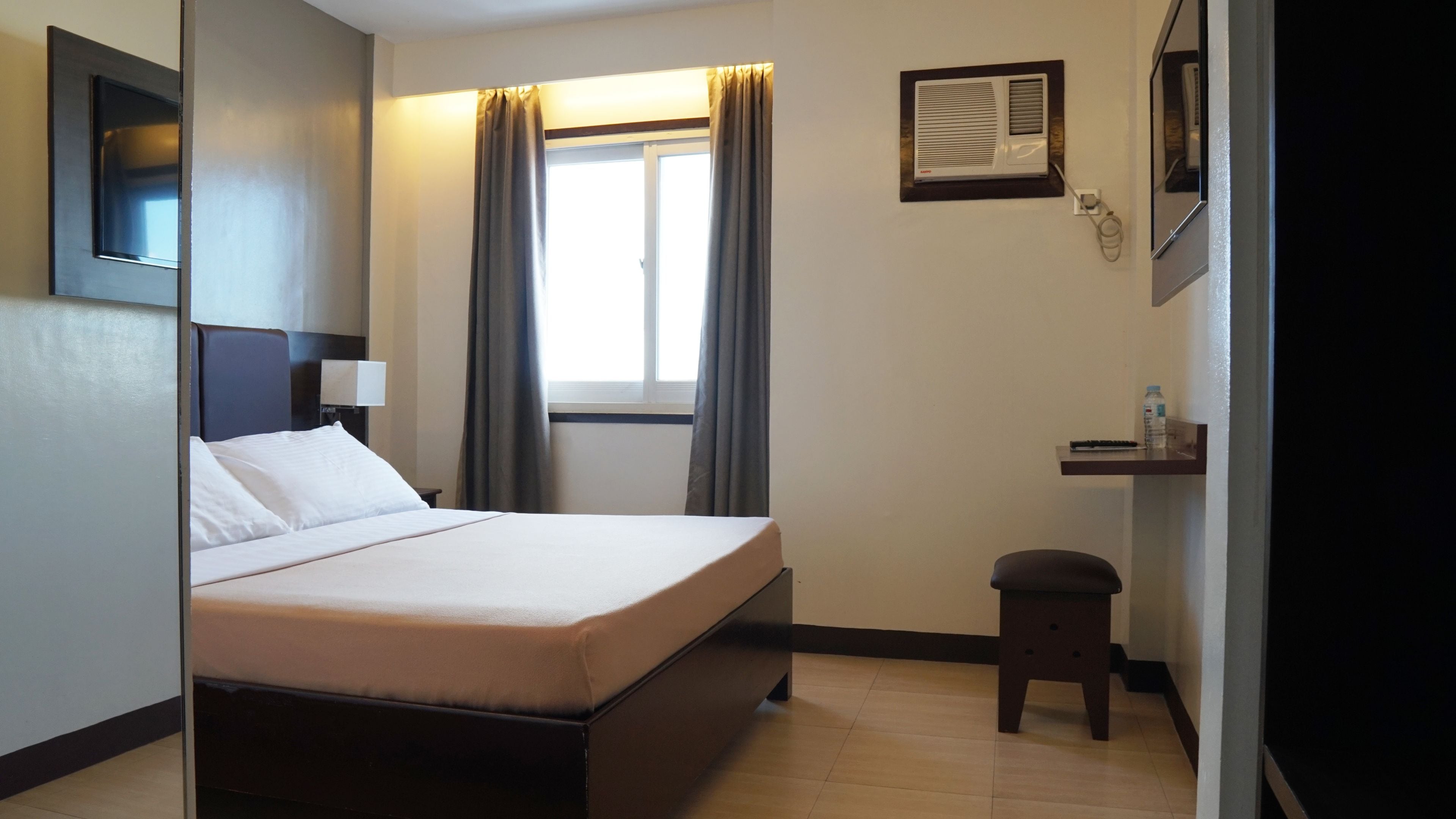 A hotel room with a bed, a nightstand, a small table with a bottle, a mirror, and a window with curtains.
