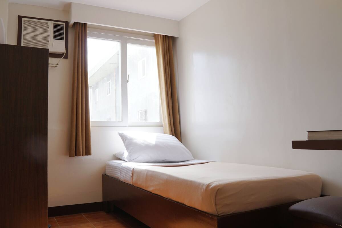 Un dormitorio pequeño y ordenado con una cama individual, una ventana con cortinas, un armario de madera y un equipo de aire acondicionado instalado en la pared.