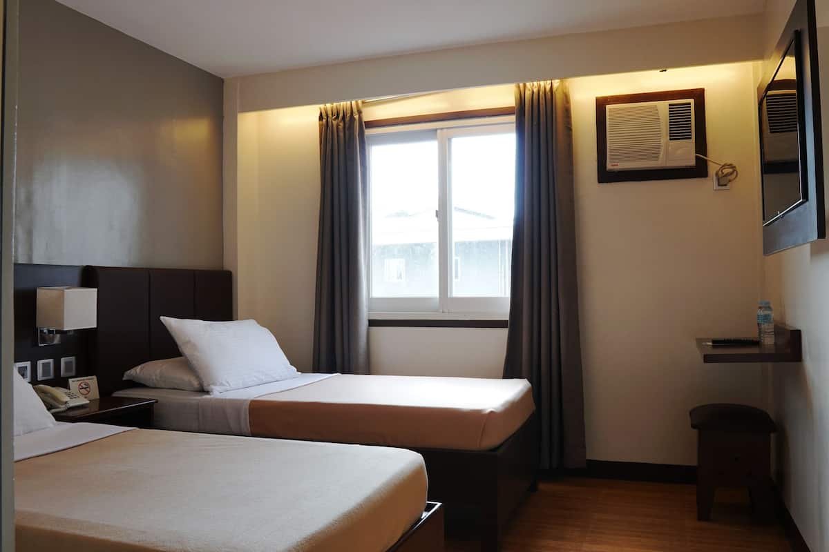 Habitación de hotel con dos camas individuales, un escritorio pequeño con silla, una mesita de noche con lámpara, una ventana con cortinas, un equipo de aire acondicionado y una botella de agua sobre el escritorio.