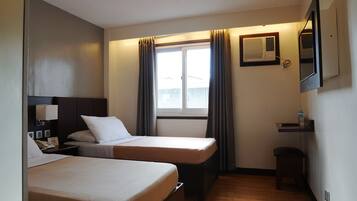 Hotelkamer met twee eenpersoonsbedden, een klein bureau met stoel, een nachtkastje met lamp, een raam met gordijnen, een airconditioner en een waterfles op het bureau.