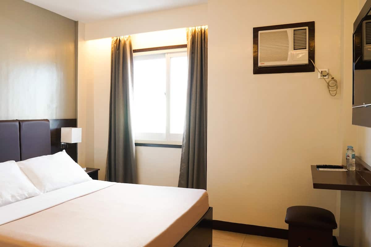 Habitación de hotel con una cama grande, una mesita de noche con lámpara, un escritorio con silla, una ventana con cortinas y un equipo de aire acondicionado.