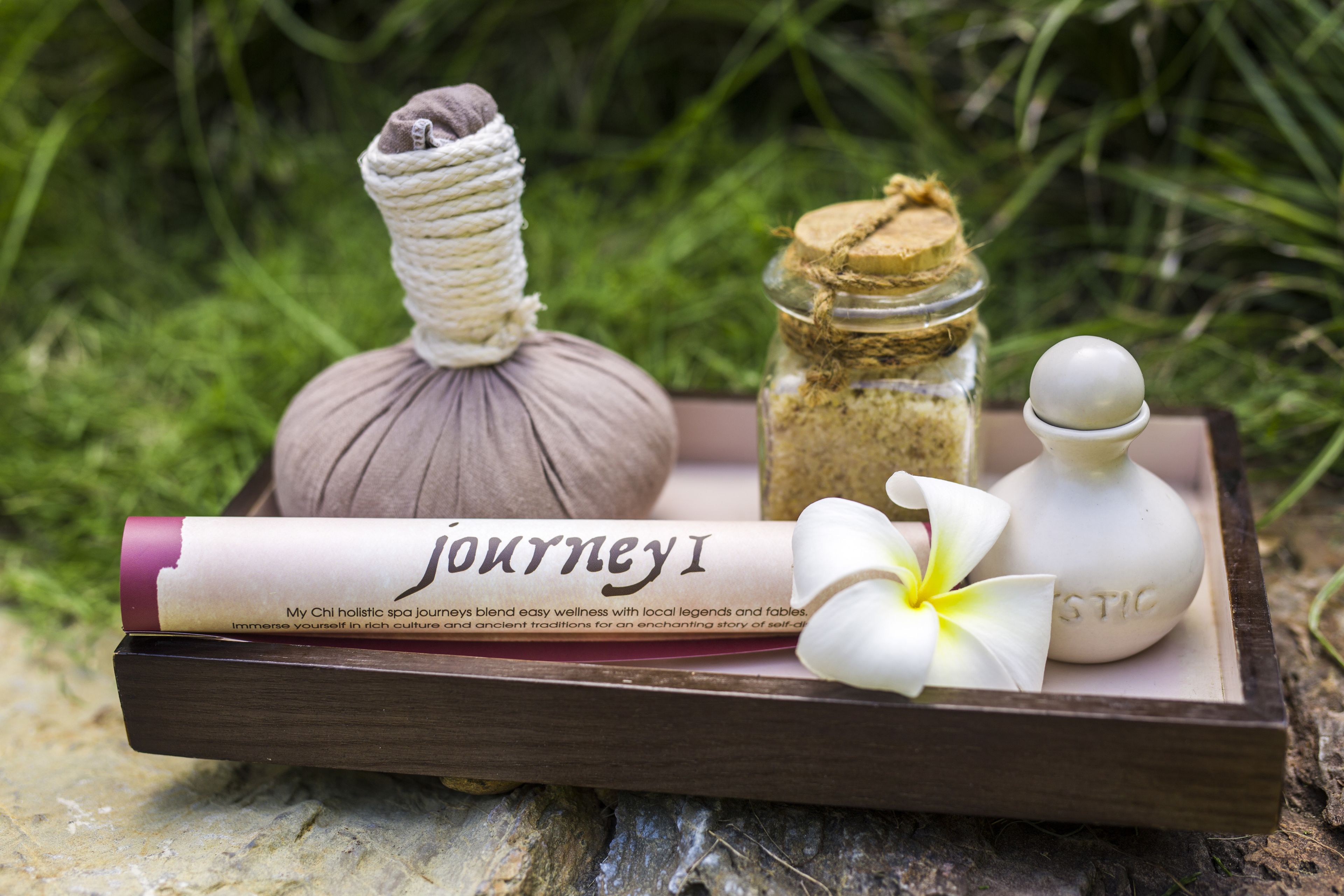 A wooden tray with a rolled-up booklet, a massage stone, a small jar with a lid, and a white bottle with a round stopper.
