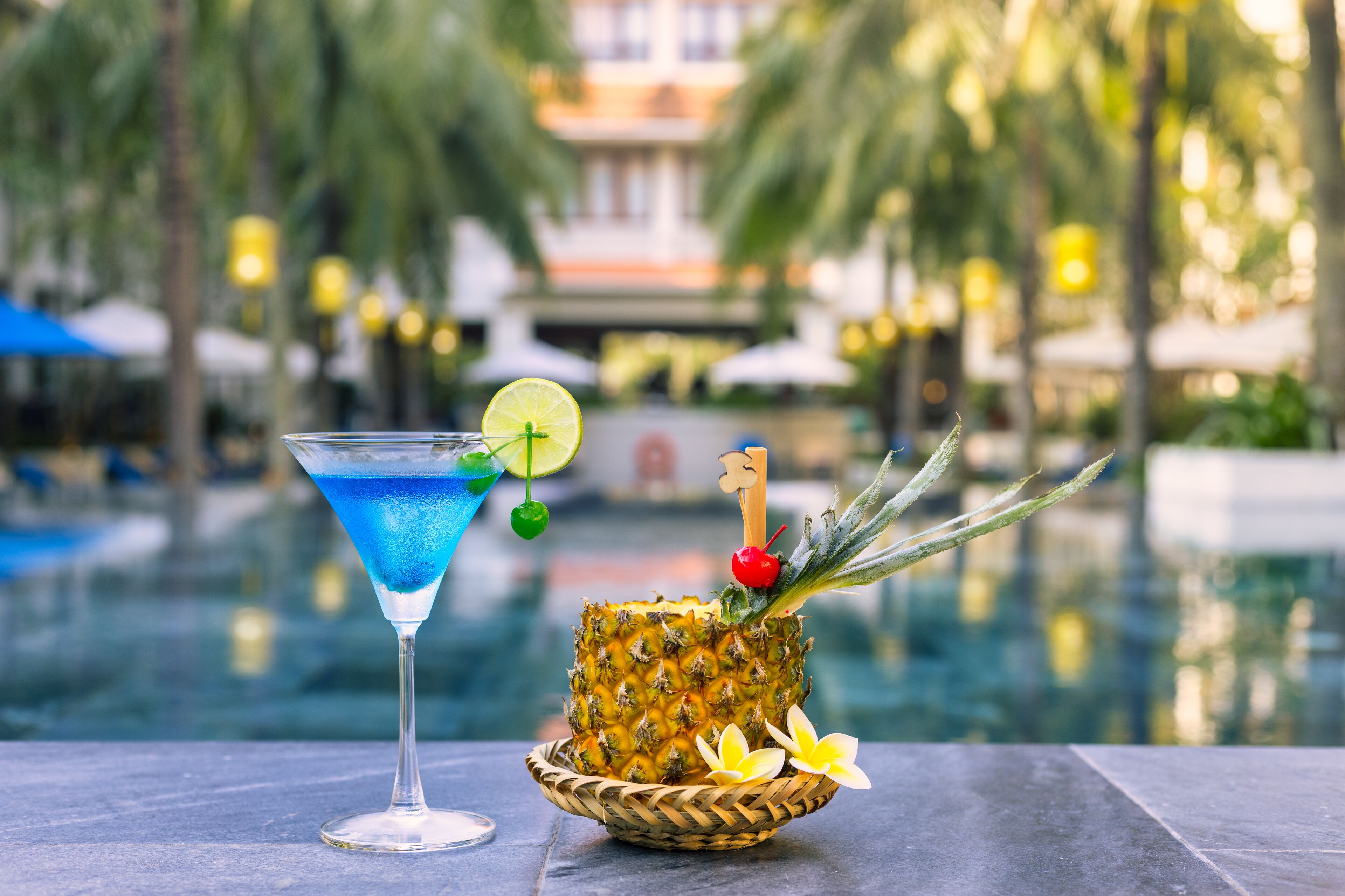 A tropical drink and a pineapple with a decorative stick, set against a poolside view with palm trees and a building in the background.