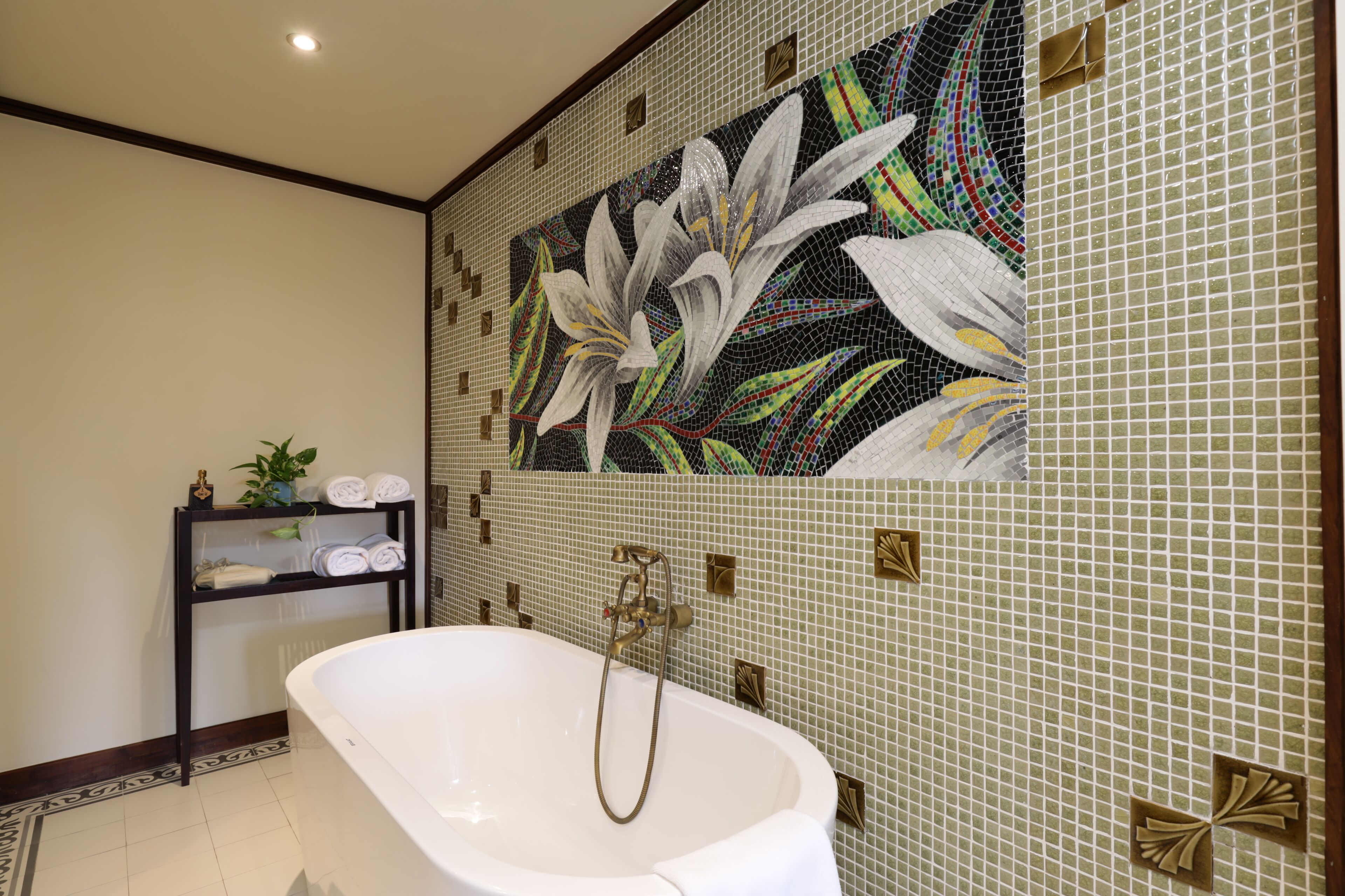A bathroom with a freestanding bathtub, a tiled wall with a floral mosaic, and a shelf with towels and a plant.