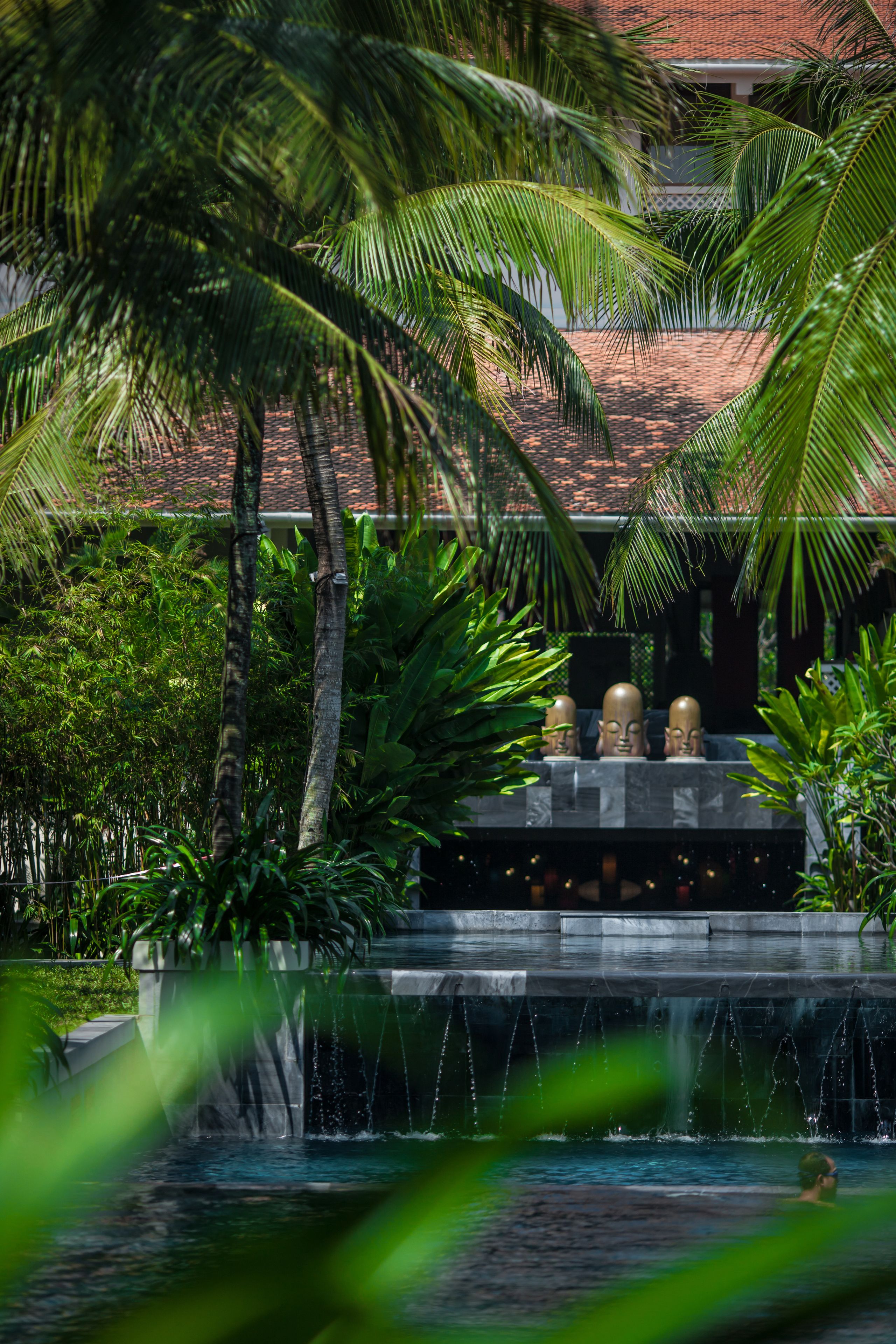 A water feature with three statues, surrounded by lush greenery and a building with a tiled roof.