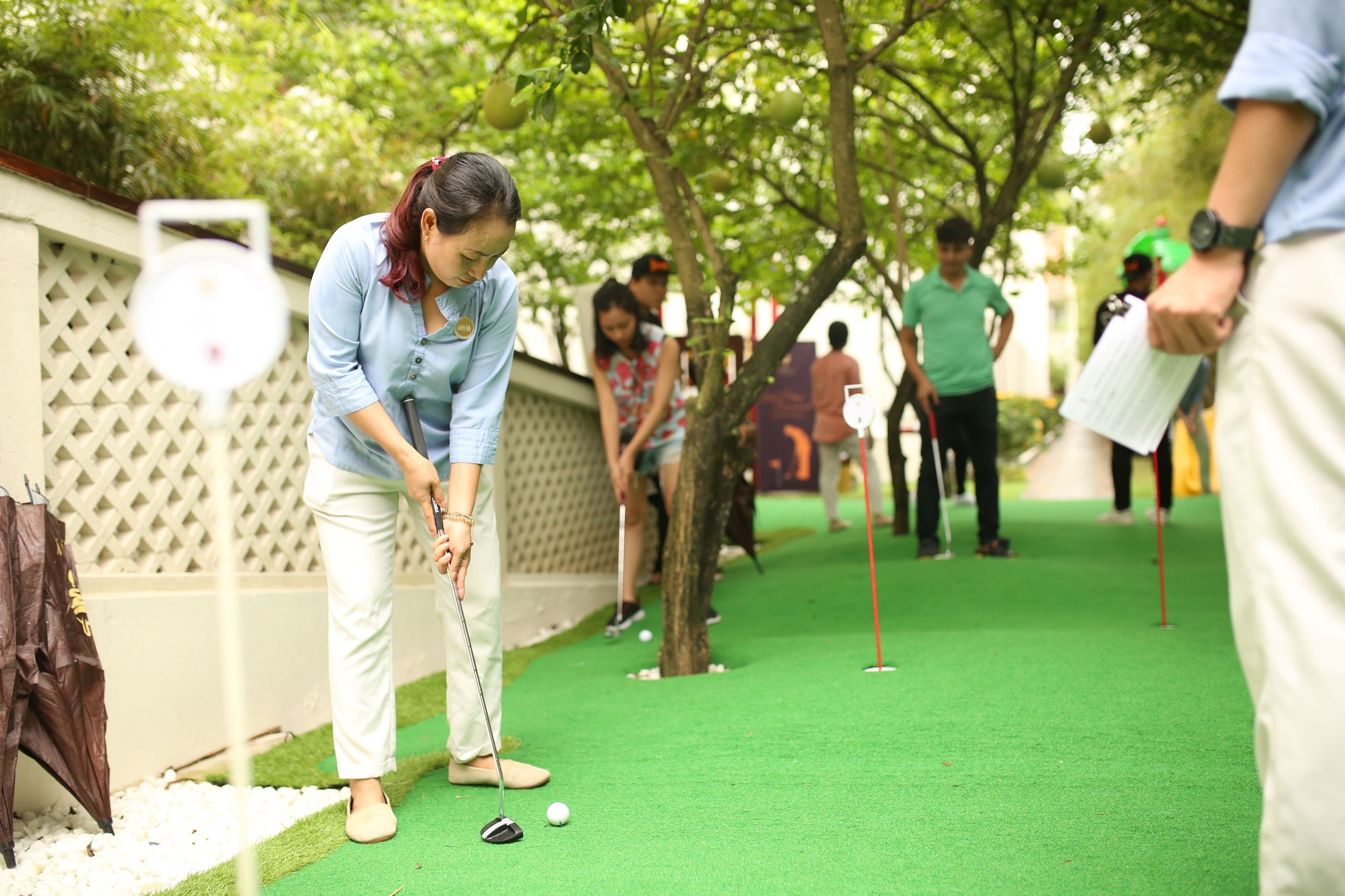A person playing golf on a green artificial turf with trees and a fence in the background.