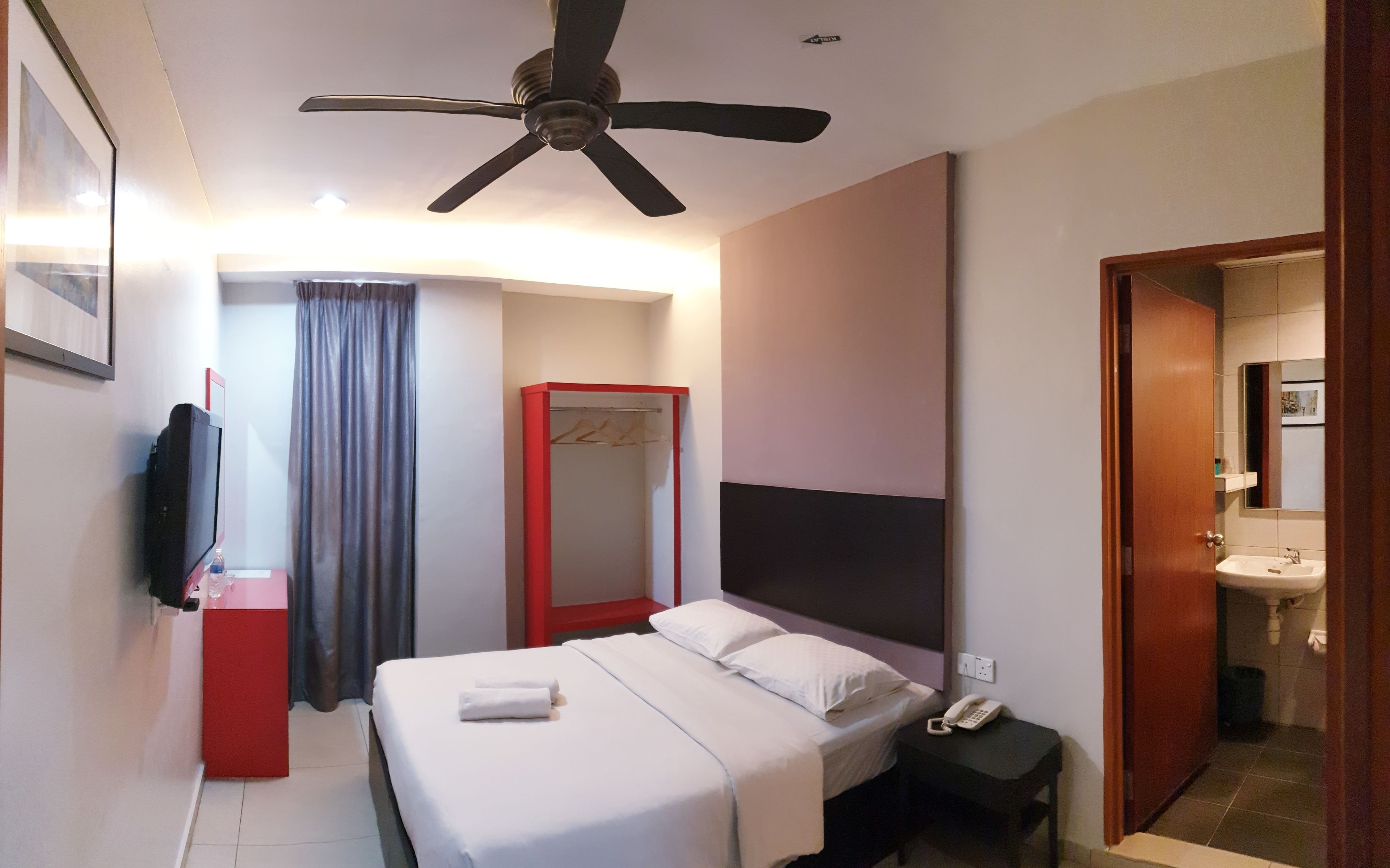 A hotel room with a bed, a bedside table with a phone, a ceiling fan, a red-framed wardrobe, a bathroom with a sink and mirror, and a TV on the left side.