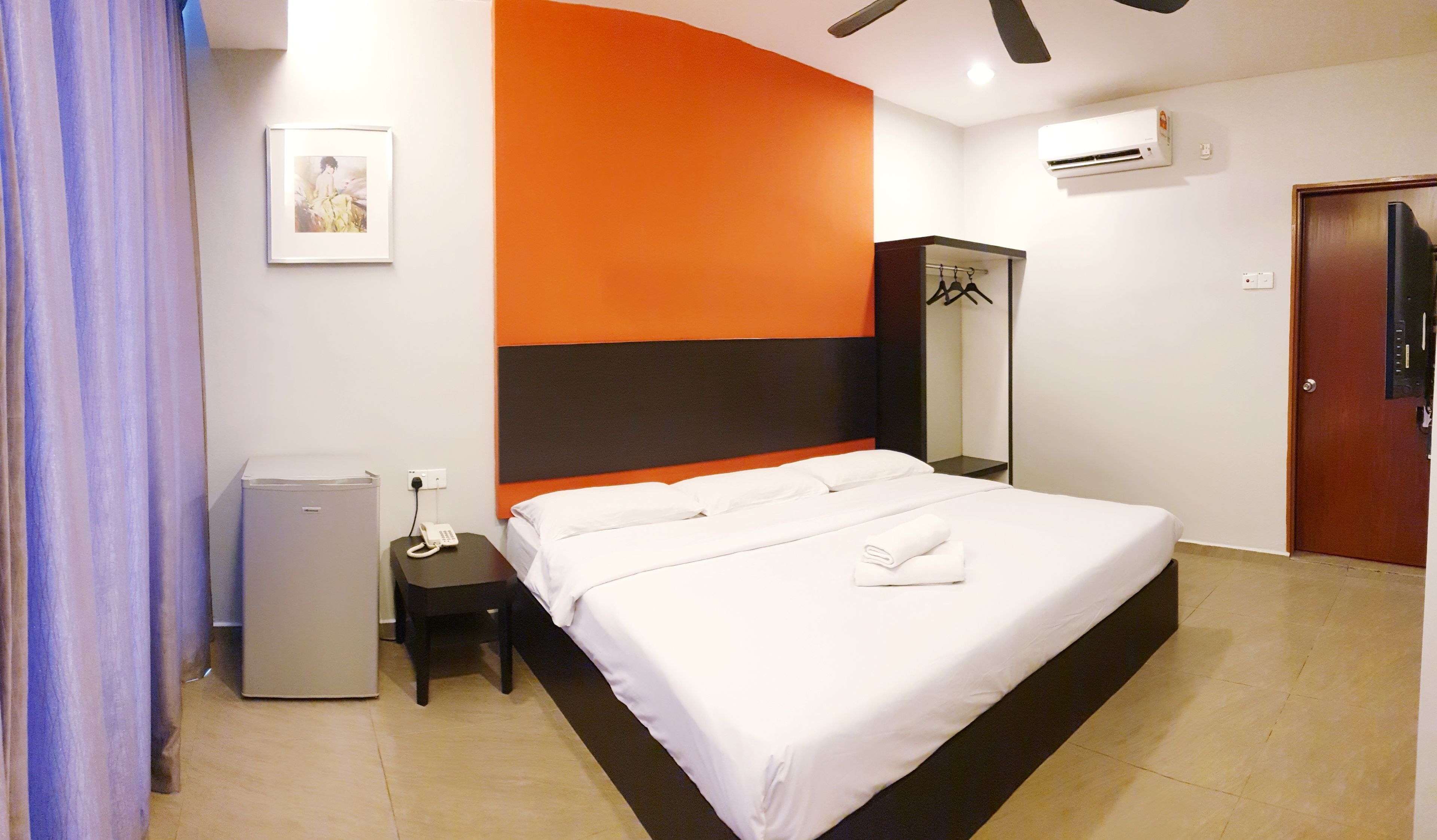 A hotel room with a bed, a bedside table with a phone, a small fridge, a mirror, and a door.