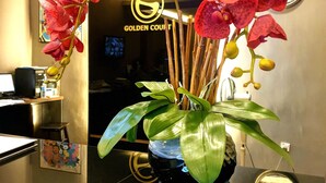 Uma mesa de recepção com um vaso de orquídeas vermelhas, um relógio e uma placa com a inscrição 'GOLDEN COURT'.