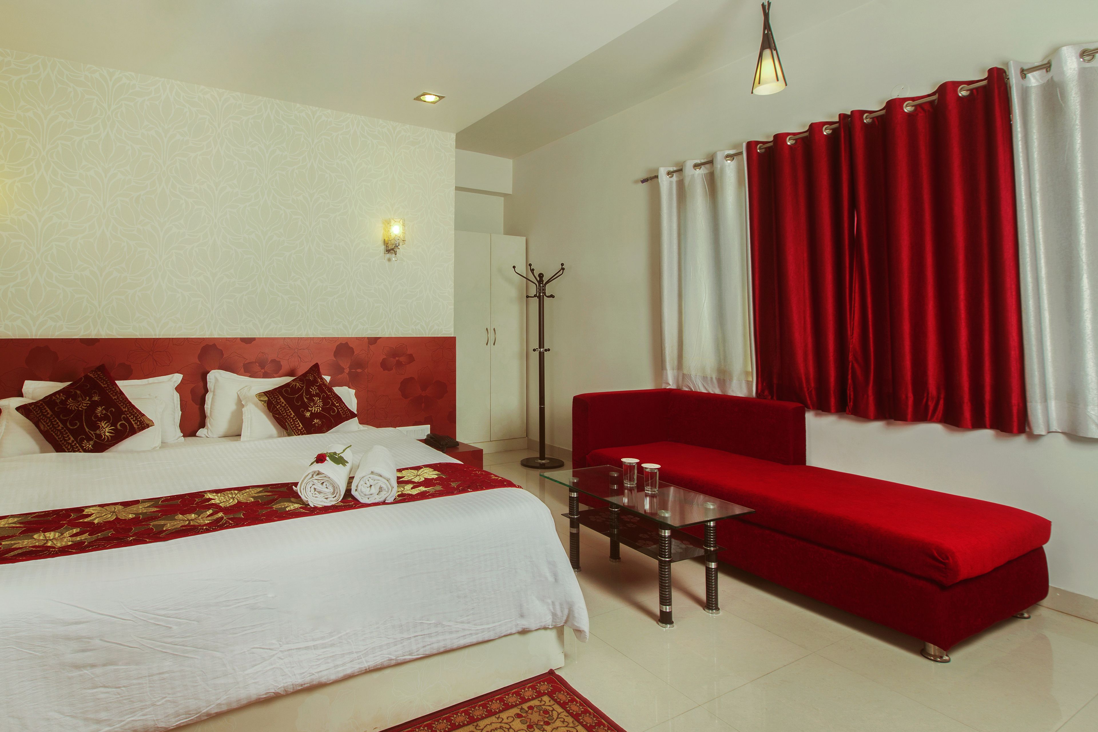 Habitación de hotel con cama, sofá rojo, mesa de centro de vidrio y cortina roja y blanca.