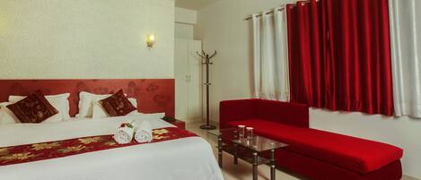 Habitación de hotel con cama, sofá rojo, mesa de centro de vidrio y cortina roja y blanca.