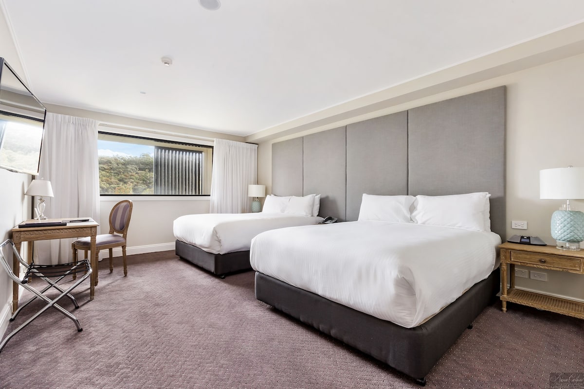 A hotel room with two beds, a desk, a chair, and a window with curtains.