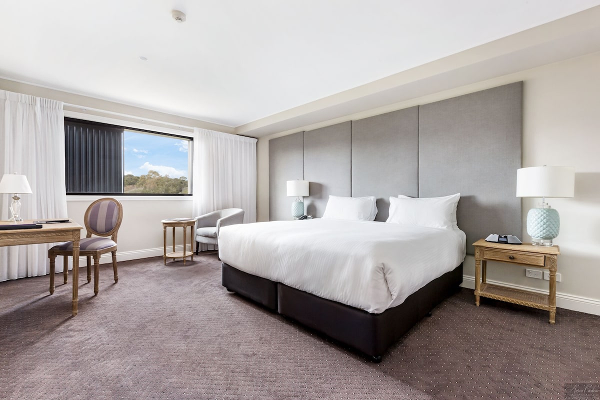 A hotel room with a large bed, two bedside tables with lamps, a desk with a chair, and a view of the outdoors through a window.
