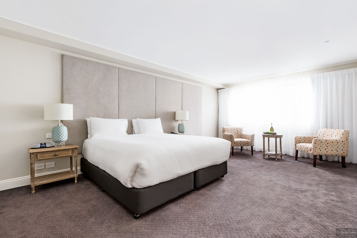 A spacious hotel room with a large bed, two bedside tables, a small table, and two chairs.
