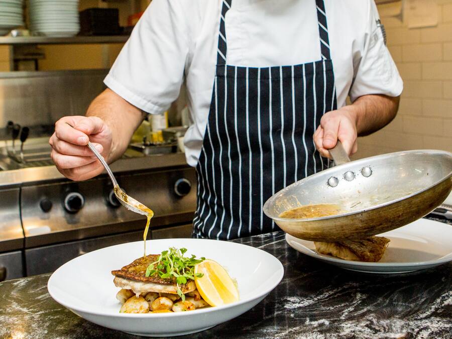 A chef preparing a dish with a frying pan and a bowl on a marble countertop.