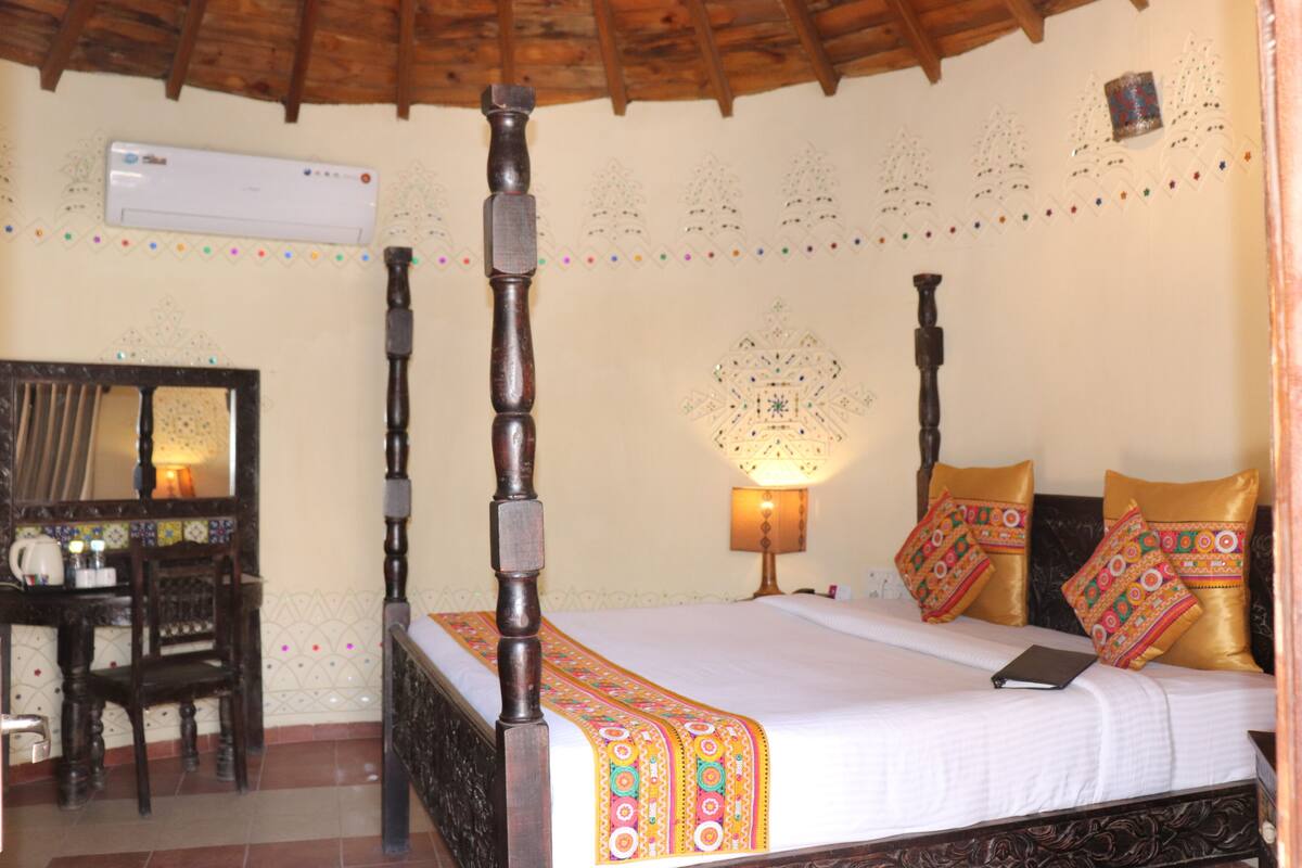 A four-poster bed with a white bedspread and colorful pillows, a wooden desk with a chair, and a wall-mounted air conditioner.