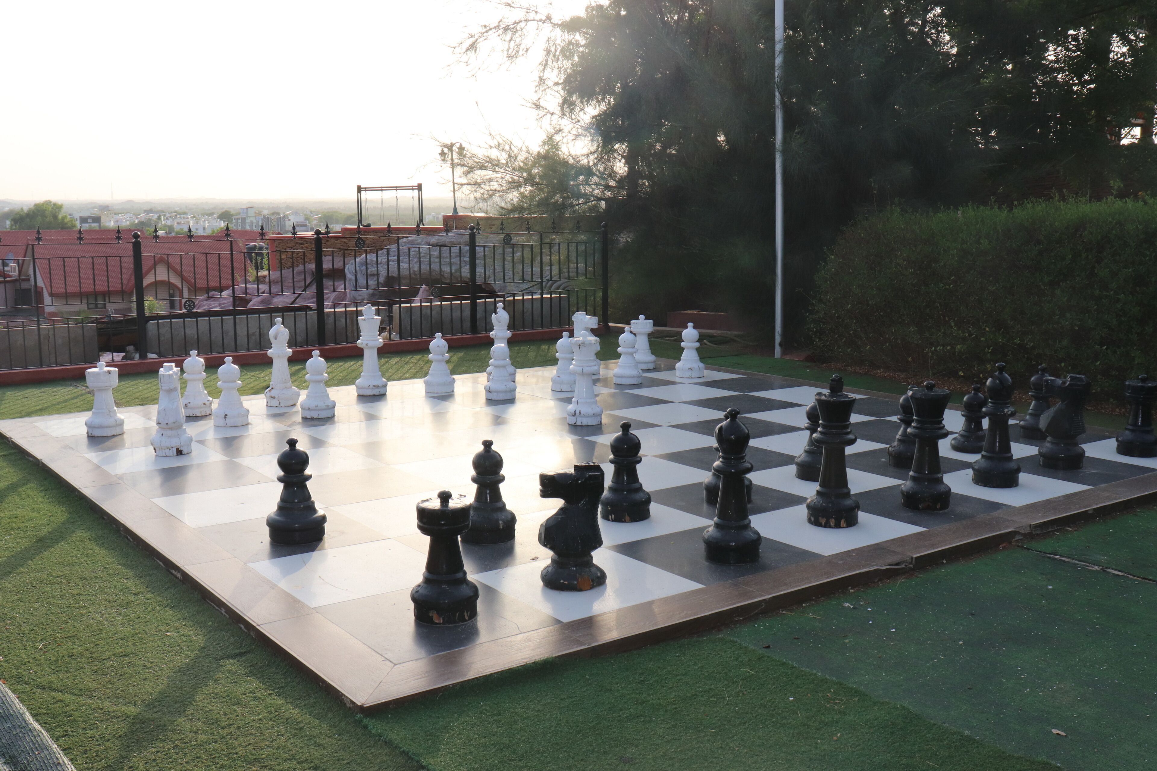 A large outdoor chessboard set up on a grassy area with a fence and trees in the background.
