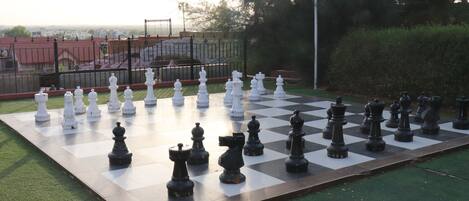 A large outdoor chessboard set up on a grassy area with a fence and trees in the background.