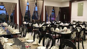Un comedor con varias mesas dispuestas para el servicio, con sillas negras de respaldo decorativo y amplias ventanas con cortinas.