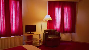 Una habitación de hotel con una cama, un televisor sobre un mueble, un sillón de cuero y cortinas rojas.