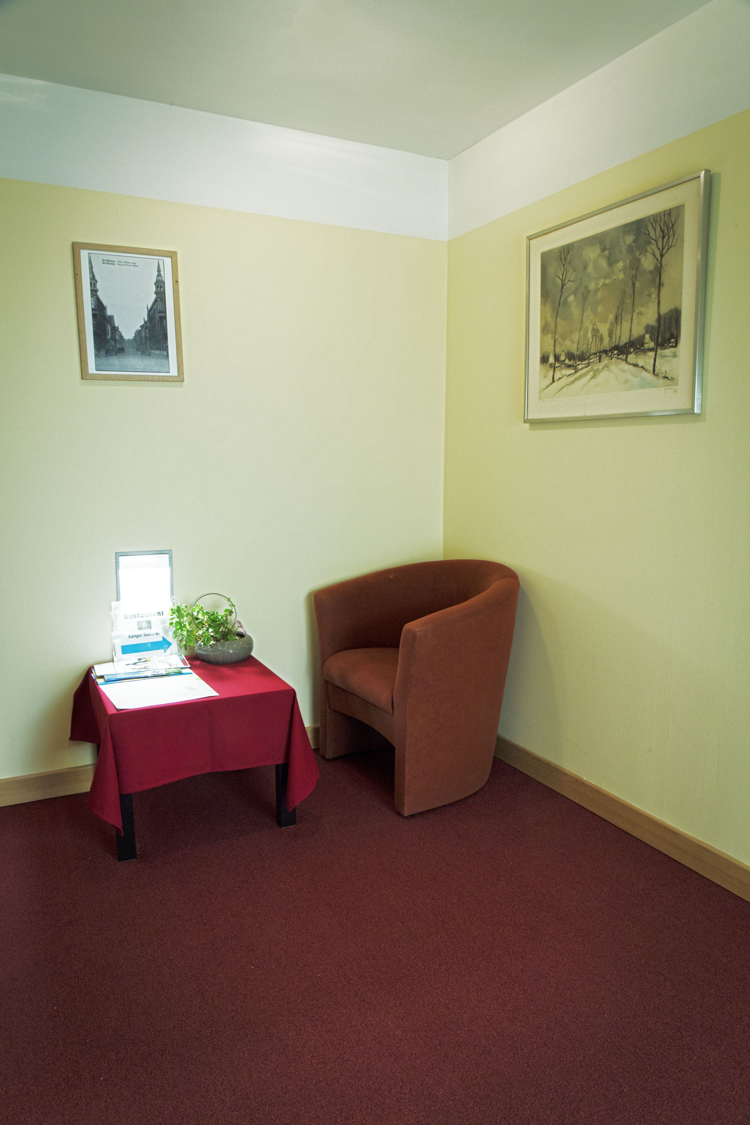 A corner room with a red carpet, a brown armchair, a small table with a plant, and two framed pictures on the walls.
