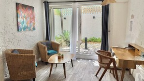 A room with wicker furniture, a small table, and a view through a glass door to an outdoor area with plants and a patterned wall. - KOHO-SXM (Grand Case)