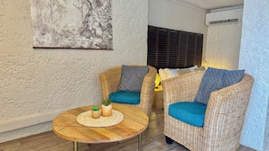 A room with two wicker chairs with blue cushions, a round wooden table with a woven placemat and a small potted plant, and a wall-mounted air conditioning unit. - KOHO-SXM (Grand Case)