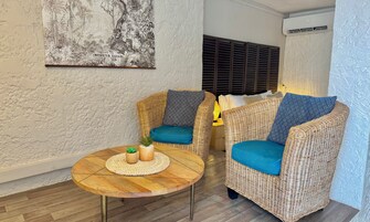 Una habitación con dos sillones de mimbre con cojines azules, una mesa redonda de madera con un mantel tejido y una pequeña planta en maceta, además de un aire acondicionado instalado en la pared.