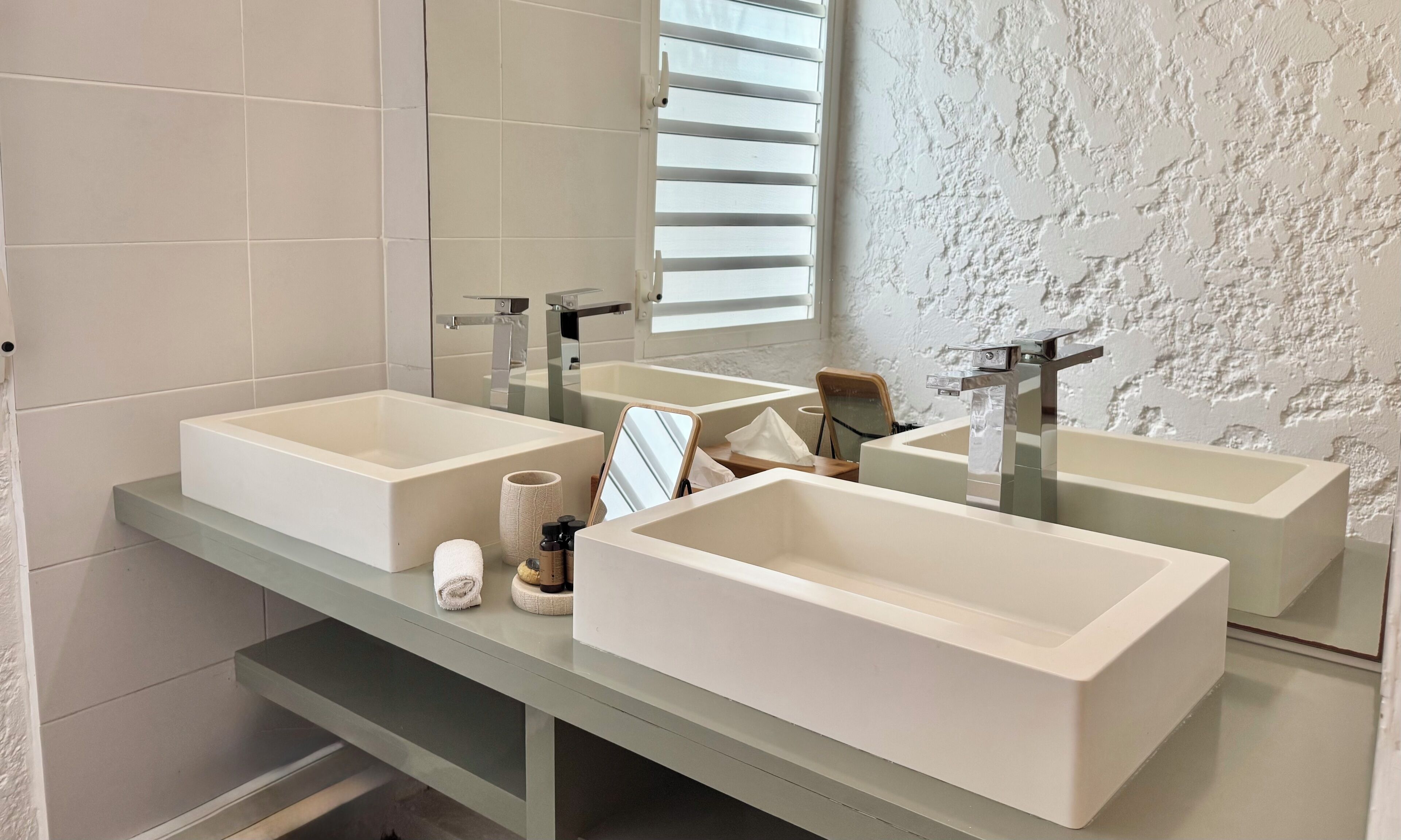 Un baño con dos lavamanos grandes de color blanco, un espejo y una ventana con persianas.