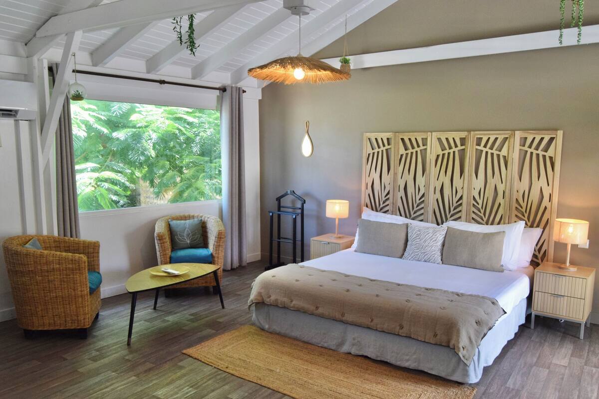 A bedroom with a large bed, two nightstands, a chair, a small table, and a window with a view of greenery.