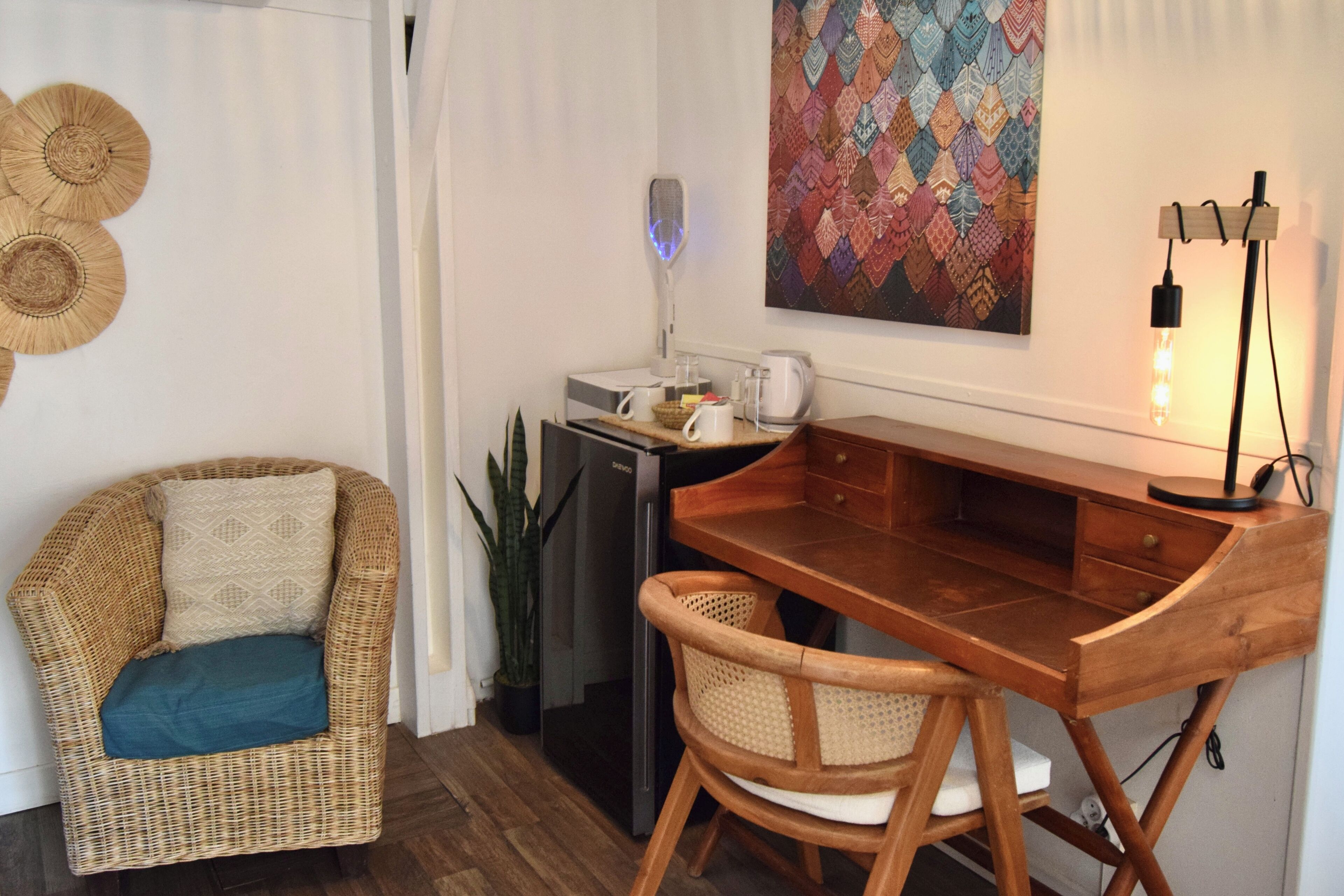 A cozy room with a wicker chair, a wooden desk, a coffee maker, and a decorative wall piece.