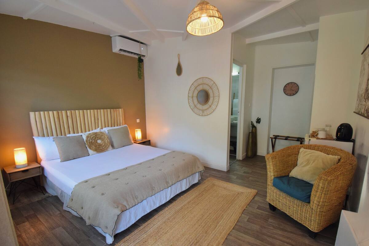 A bedroom with a bed, bedside tables, a wicker armchair, a wall-mounted air conditioner, and a hanging light fixture.