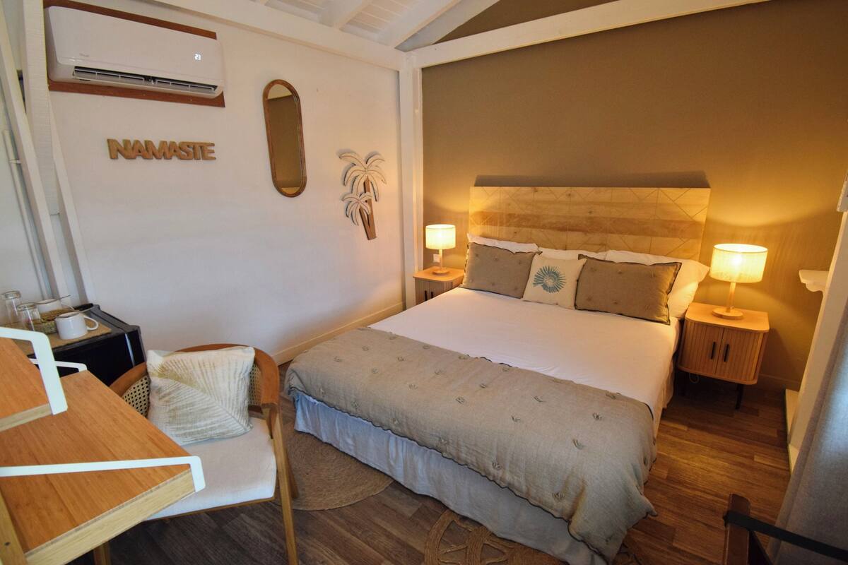 A neatly arranged bedroom with a large bed, two bedside tables with lamps, a chair, a small table with a cup, and a wall-mounted air conditioner.